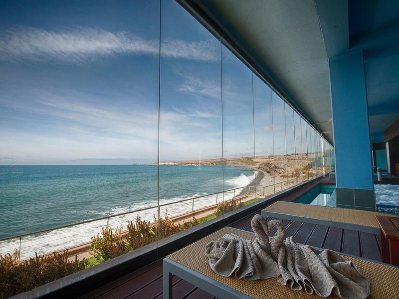 Balcón con mesa y toalla servida, vista al mar y costa bajo cielo despejado.