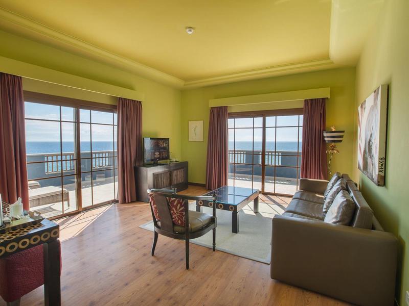 Sala luminosa con paredes verdes, sofás, sillones y vista a terraza y mar.