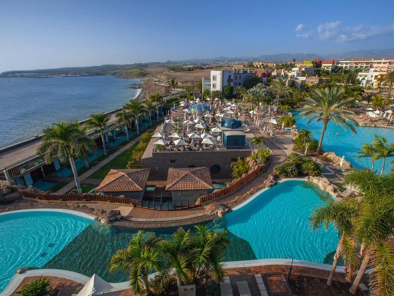 Gran piscina de hotel con palmeras y sombrillas junto al mar bajo cielo despejado.