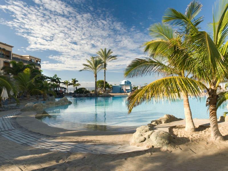 Una área de piscina tranquila con palmeras y edificios al fondo bajo un cielo azul.