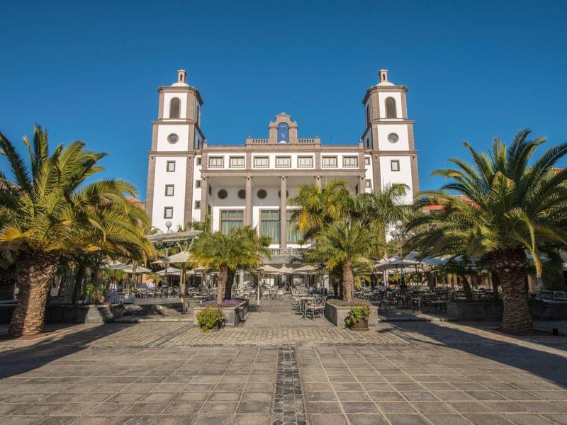 Edificio histórico grande con dos torres y palmeras en plaza adoquinada bajo cielo despejado.
