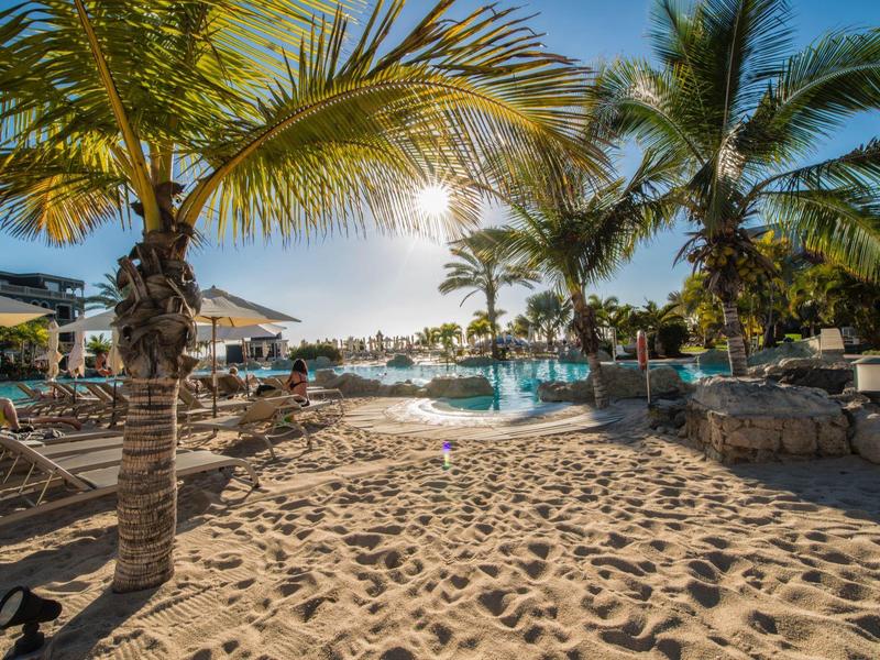 Área de playa soleada con palmeras, piscina y tumbonas en un resort hotelero tropical.
