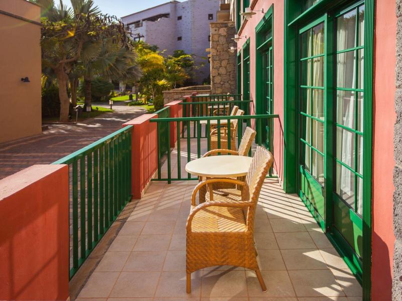 Balcón con mesa y sillas frente a ventanas verdes en un día soleado.