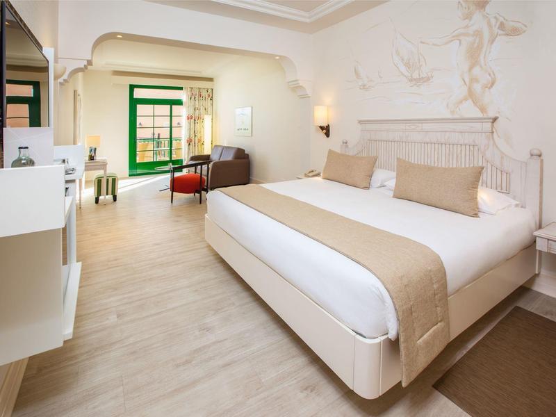 Habitación de hotel luminosa con cama grande, zona de estar y balcón con vista al patio interior.