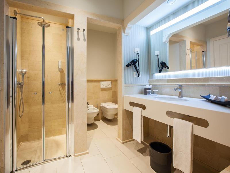 Moderno baño de hotel con ducha a ras de suelo, doble lavabo y grandes espejos en tonos cálidos.