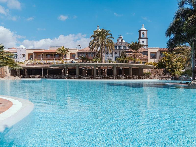 Gran piscina frente a un hotel con palmeras y cielo azul de fondo.