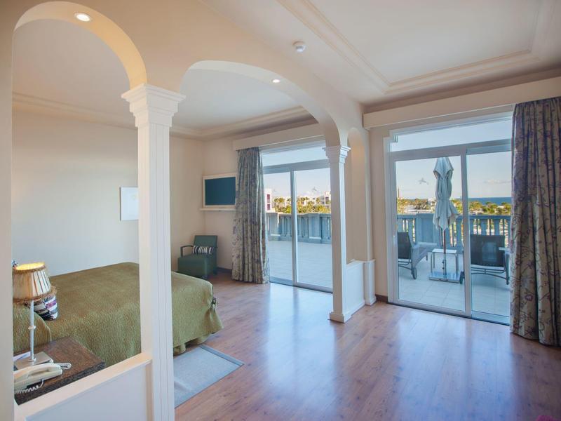 Habitación de hotel luminosa con balcón, suelo de madera y vistas al agua al atardecer.