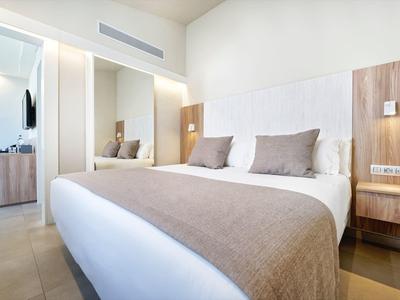 Helles Schlafzimmer mit großem Bett, hellbraunem Bettüberwurf, Kissen und modernem Holzmöbel.