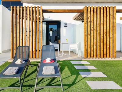 Moderne Terrasse mit weißen Wänden, Holzlamellen, grüner Wiese und zwei schwarzen Liegestühlen.