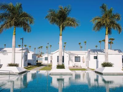 Bungalow bianchi con palme riflesse in una piscina azzurra sotto un cielo soleggiato.