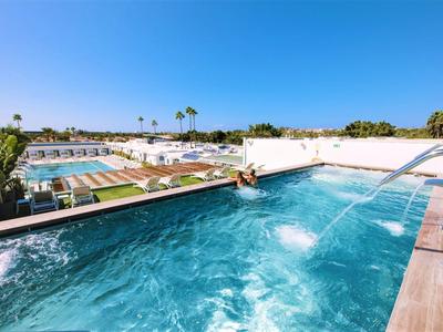 Klares Wasser im Pool mit Wasserspielen, darunter Liegewiesen und weiße Gebäude bei blauem Himmel.
