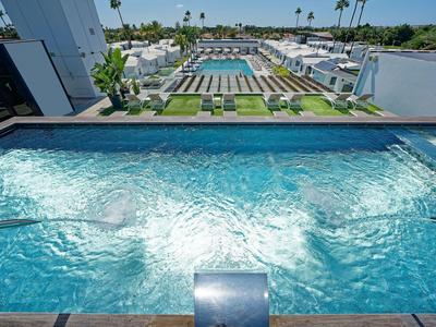 Moderner Außenwhirlpool mit Wasserstrahlen vor einem großen Schwimmbecken und Palmen im Hintergrund.