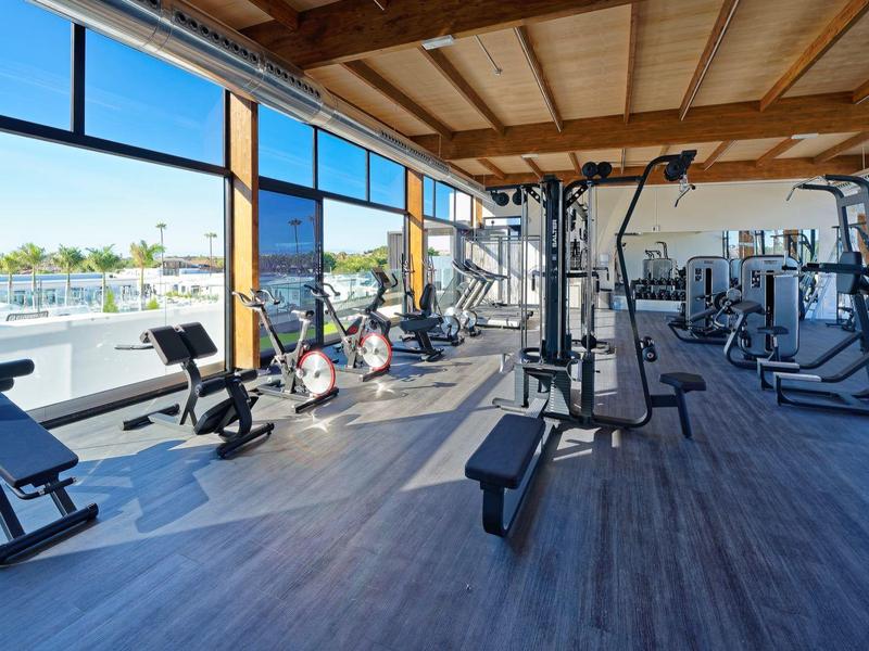 Helles Fitnessstudio mit großen Fenstern, Geräten und Holzbalkendecke, Blick auf Wasser und Bäume.