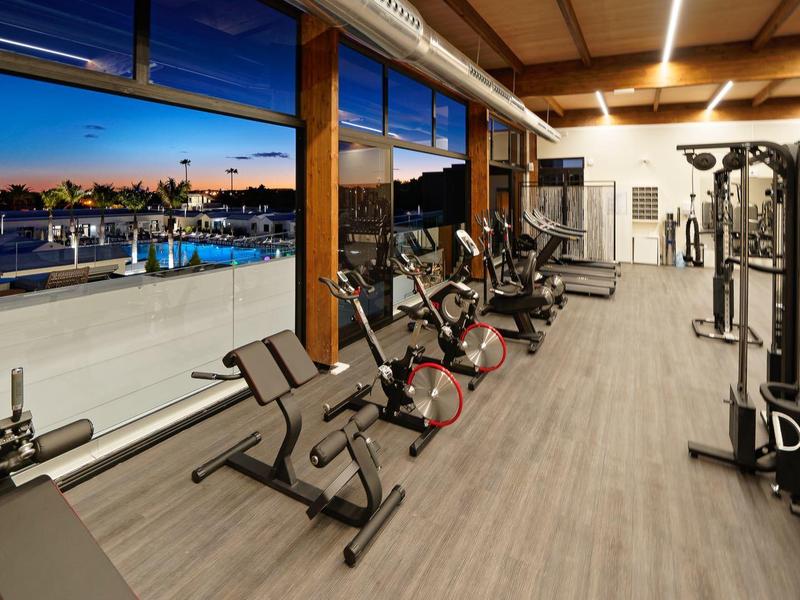 Moderner Fitnessraum mit Kraftgeräten, Hanteln und weitem Fenster mit Blick auf Pool bei Sonnenuntergang.