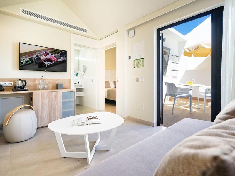 Modern eingerichtetes Wohnzimmer mit hellen Möbeln, TV, rundem Tisch und Zugang zur Terrasse.