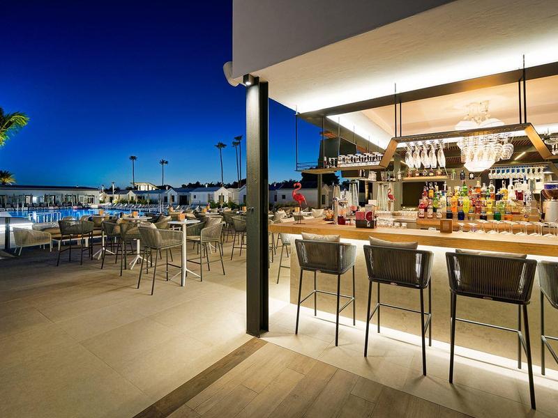 Moderner Außenbarbereich mit Hockern, dekorativen Pflanzen und Abendlicht am Wasserufer.