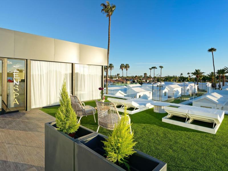 Moderne Dachterrasse mit Liegestühlen, Glasgeländer, Pflanzen in Kästen und Palmen im Hintergrund.