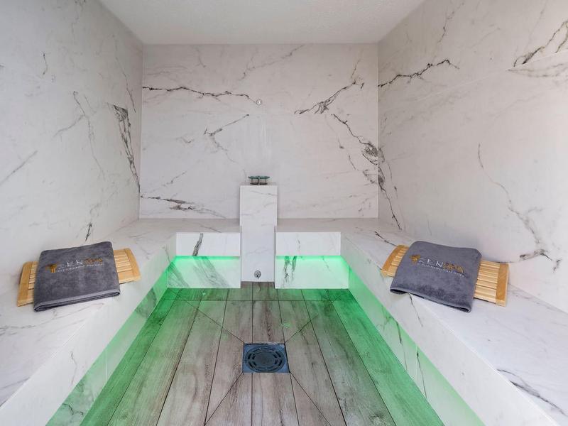 Moderner Dampfraum mit weißem Marmor, grüner LED-Beleuchtung und Holzbänken mit grauen Kissen.