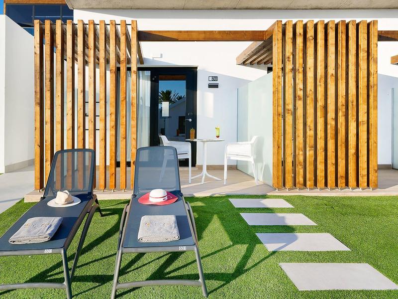 Moderne Terrasse mit weißen Wänden, Holzlamellen, grüner Wiese und zwei schwarzen Liegestühlen.