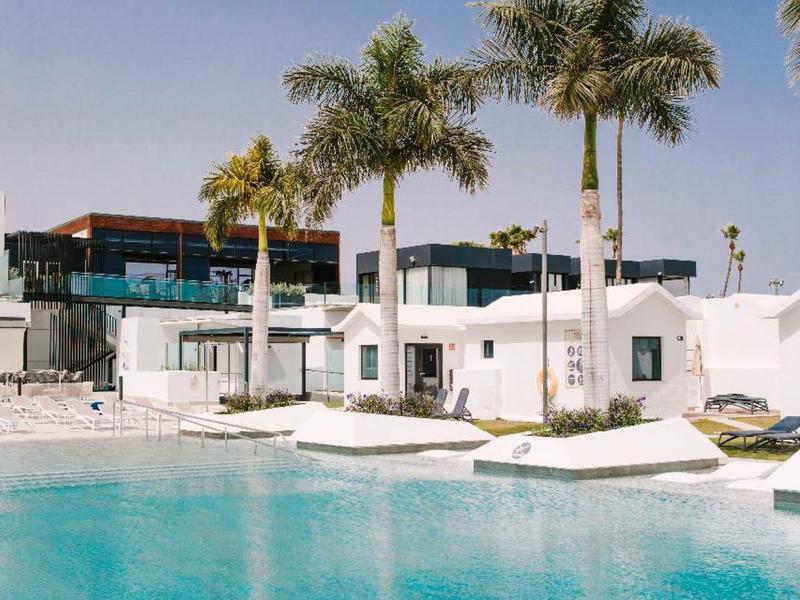 Hellblauer Pool vor weißem Gebäude mit Palmen und klarem Himmel im Hintergrund.