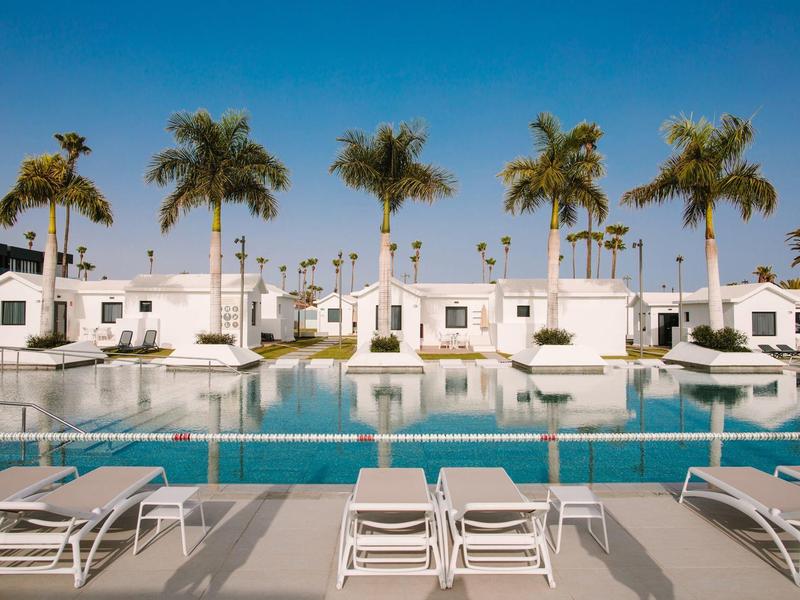 Schwimmbad mit blauer Wasserfläche, weißen Liegestühlen und Palmen vor weißen Bungalows.