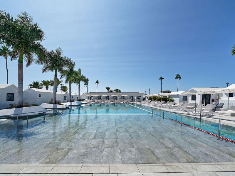 Großer klarer Pool mit Palmen und weißen Liegen unter blauem Himmel in einer Hotelanlage.