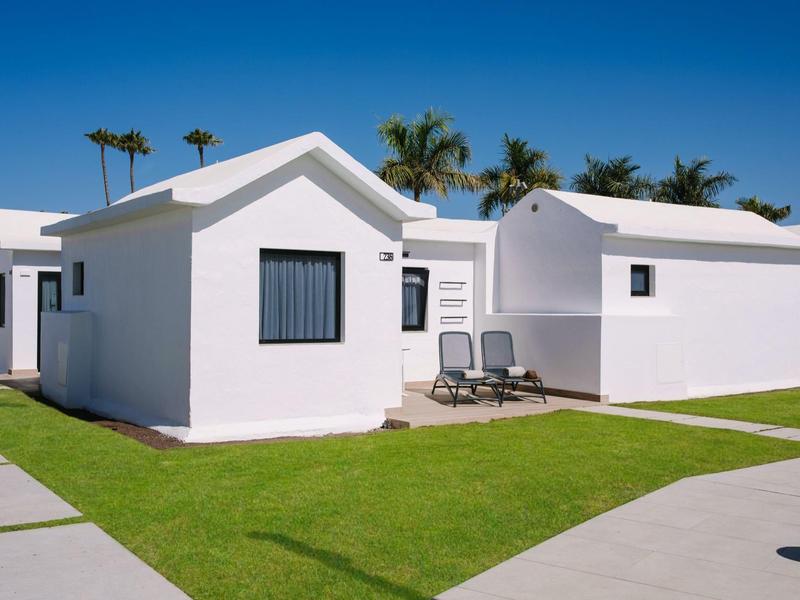 Weiße Bungalows mit Flachdach auf grünem Rasen unter blauem Himmel, davor zwei Liegestühle.