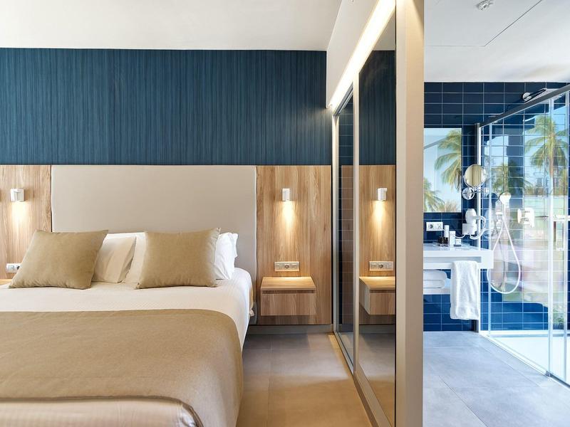 Modernes Hotelzimmer mit großem Bett, Holzkopfteil, blauer Akzentwand und bodentiefen Fenstern.