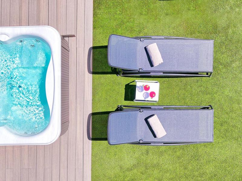 Zwei graue Liegen mit Kissen, ein kleines Tablett mit Getränken, grüner Rasen und weißer Whirlpool.