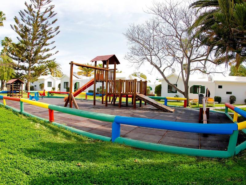 Kinderspielplatz mit buntem Holzzaun, Rutsche und Klettergerüst, umgeben von grünem Gras.