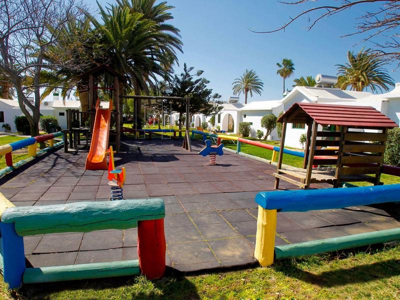 Kinderspielplatz mit Rutsche, Schaukeln und buntem Zaun aus Holz auf grünem Rasen bei Sonnenschein.