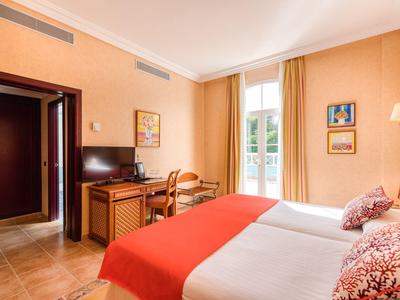 Hell eingerichtetes Hotelzimmer mit zwei Betten und orangem Bettüberwurf, Schreibtisch und Fenster.