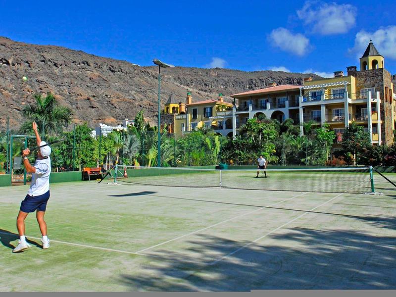 People playing tennis on a court with a hotel and mountain in the background.