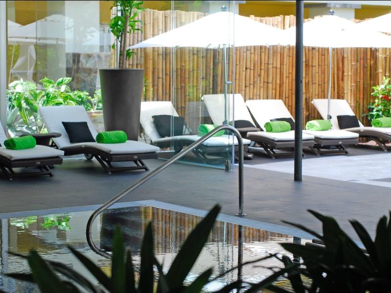 Poolside area with lounge chairs and umbrellas surrounded by plants and a wooden fence.