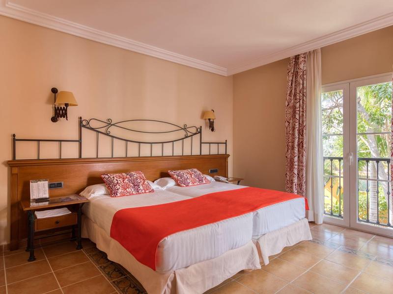 Cozy hotel room with twin beds, red accents, and a balcony overlooking greenery.