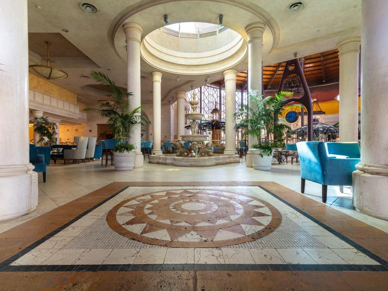Spacious hotel lobby with pillars, circular ceiling design, plants, and turquoise seating.