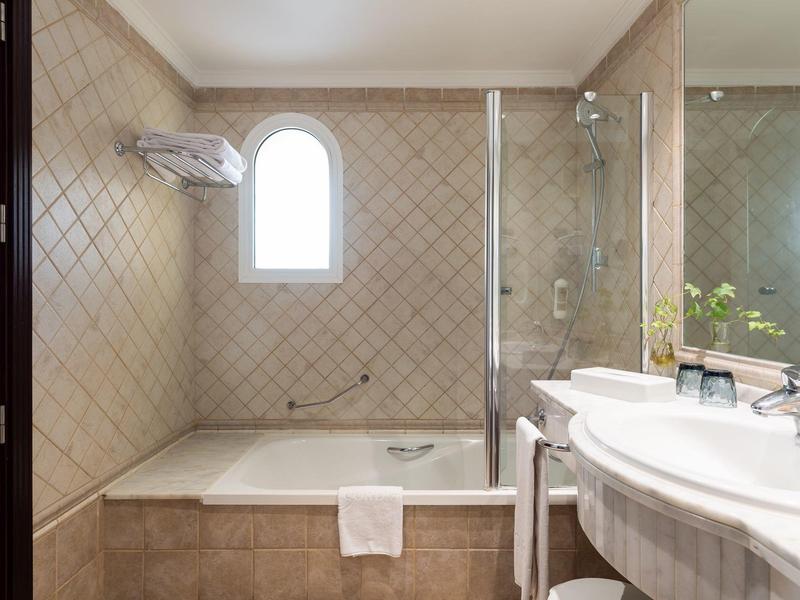 Bathroom with a bathtub, towel, and sink with a mirror, featuring beige tiled walls.
