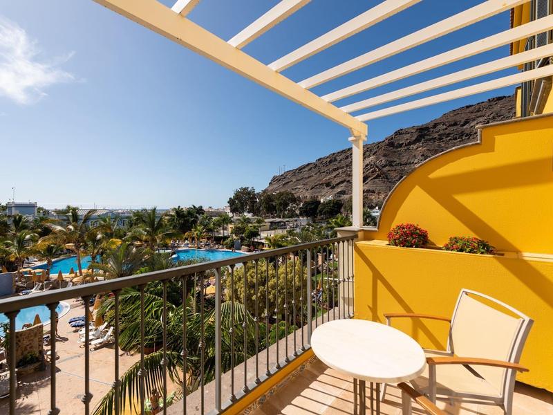 Balcony with chairs overlooks palm trees, pool, and mountain under clear blue sky.