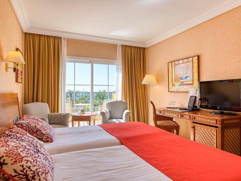 Hotel room with two beds, red throws, armchairs, desk with TV, and balcony with a view.