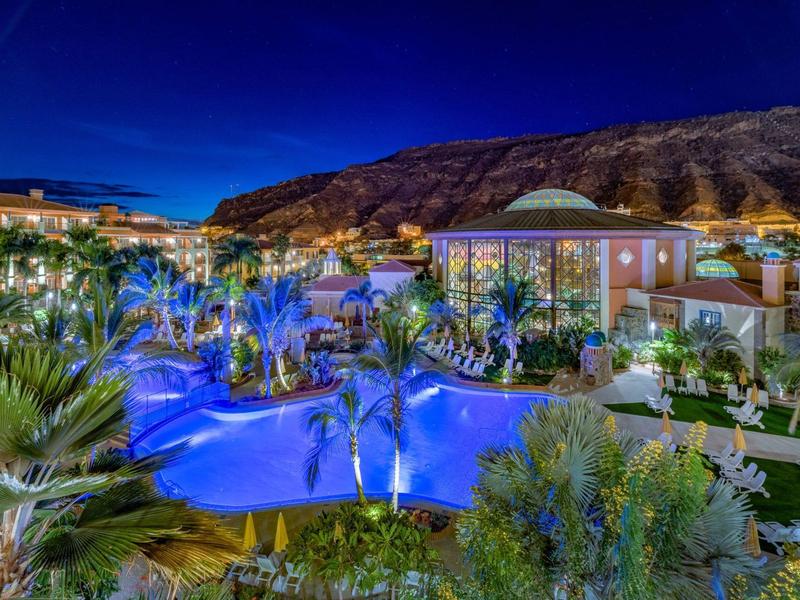 Luxury hotel pool illuminated at night with palm trees and mountain backdrop