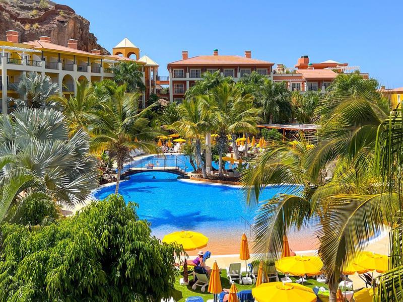 Resort with blue swimming pool surrounded by palm trees, yellow umbrellas, and buildings under clear sky.