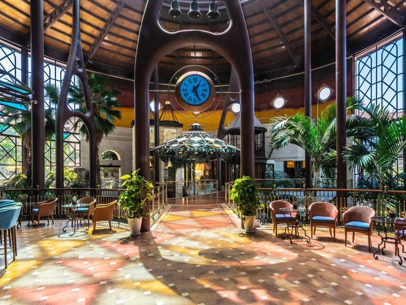 Bright hotel lobby with large windows, plants, and circular seating under a high wooden ceiling.