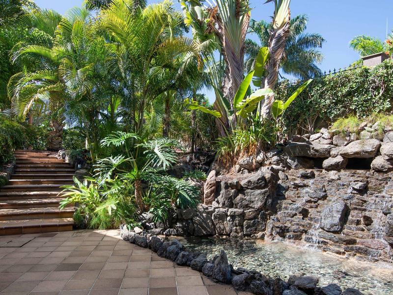 Lush garden with stone waterfall and steps leading upward under blue sky.