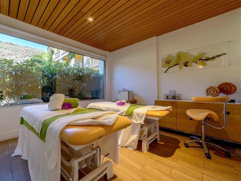 Cozy spa room with two massage tables, wooden furniture, and a large window overlooking greenery.