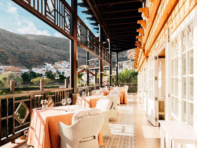 Sunny outdoor terrace with tables and chairs overlooking a mountain village.