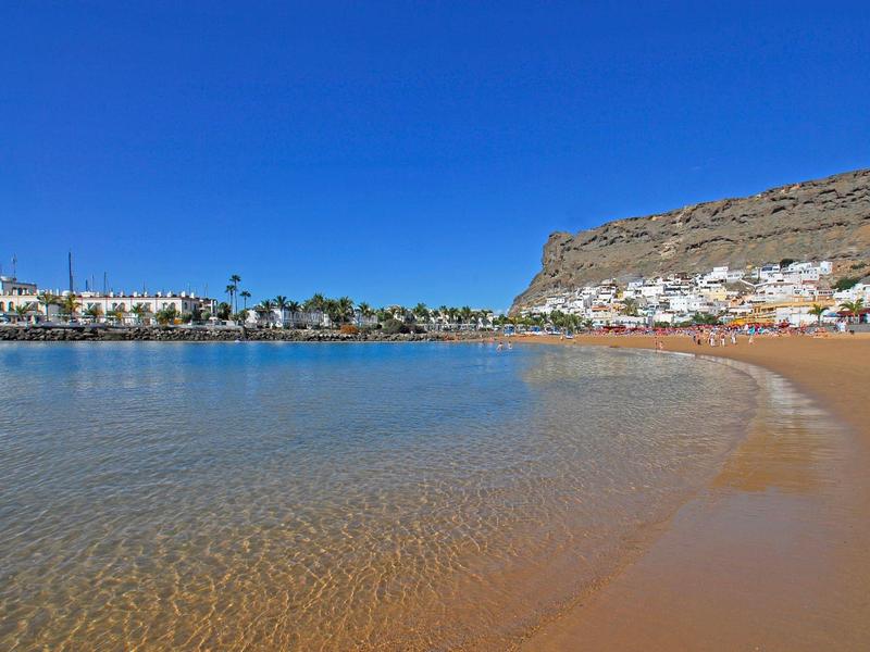 Clear beach with calm water, sandy shore, buildings, and a rocky hill under a bright blue sky.