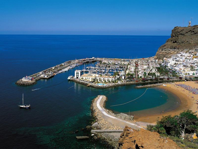 Coastal town with marina, crescent beach, and white buildings on a cliff by the sea.