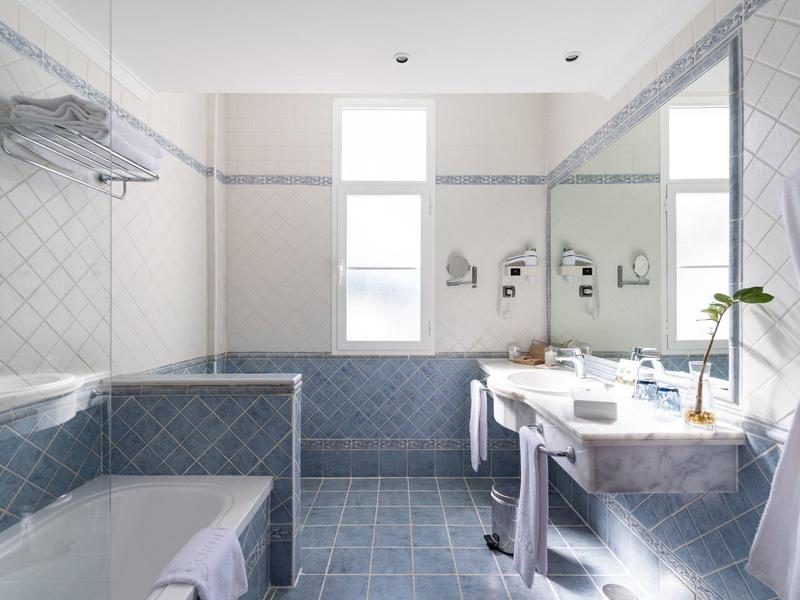 Bright bathroom with blue tiled floor, bathtub, large mirror, and twin windows.
