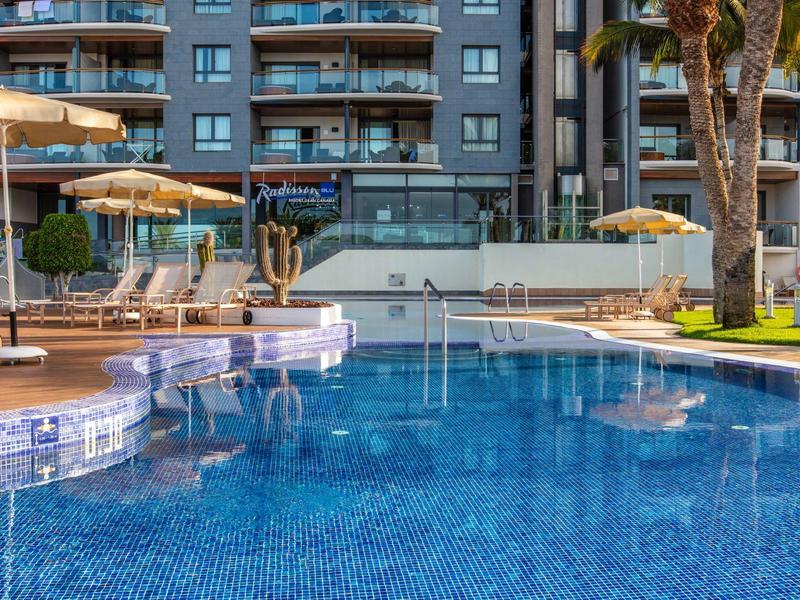 Čistý bazén s modrými mozaikovými dlaždicemi před vícepatrovým hotelem s slunečnímimi parasoly.