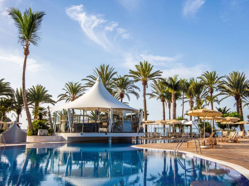 Luxusní bazénová zóna s bílými stany, palmami a modrou oblohou v rezortu.
